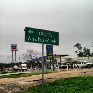 This is an image of a traffic sign near Houston, TX. It's got a dark green background with white lettering. The top line of the sign has an arrow pointing to the left, suggesting drivers turn left to head toward the city called "Liberty." Below, there is an arrow pointing to the right, informing drivers to turn right to head to a city called "Anahuac." The sign rests in front of a Chevron gas station, which you can see in the background.